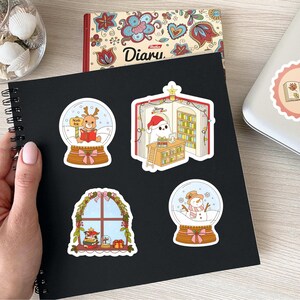 May include: A black spiral-bound notebook with four Christmas-themed stickers. The stickers feature a reindeer in a snow globe, a ghost in a Santa hat in a library, a window scene, and a snowman in a snow globe. A diary and seashells are also visible.