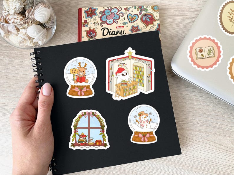 May include: A black spiral-bound notebook with four Christmas-themed stickers. The stickers feature a reindeer in a snow globe, a ghost in a Santa hat in a library, a window scene, and a snowman in a snow globe. A diary and seashells are also visible.