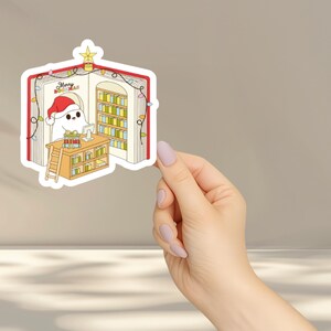 May include: A sticker featuring a cartoon ghost wearing a Santa hat, inside a book-shaped library. The ghost is at a desk with presents, and the library has bookshelves and Christmas lights. The text "Merry Bookmas" is visible.