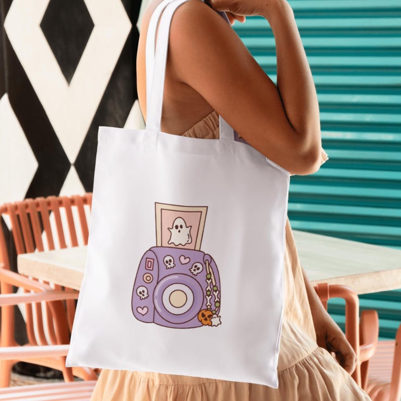 May include: White tote bag featuring a purple camera illustration with a ghost photo, skulls, and a pumpkin charm. The bag has white handles and is held by a person. The design has a Halloween theme.