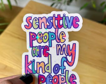 May include: A colorful sticker with the text "Sensitive people are my kind of people." The text is in various colors, including pink, blue, and purple, with a holographic glitter effect. The sticker is held by a hand.
