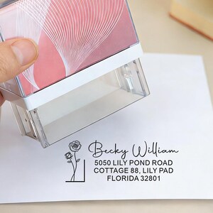 Puede incluir: Un sello de dirección transparente con una base blanca y una parte superior con estampado rosa y blanco se utiliza para estampar un sobre blanco. El texto estampado incluye un nombre, una dirección y una ilustración floral.