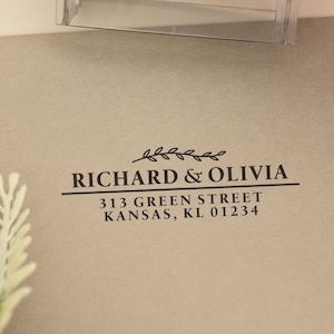 Puede incluir: Un sello transparente de la marca "COLOP" se utiliza para imprimir en un sobre marrón claro. El sobre tiene los nombres "RICHARD & OLIVIA" y los detalles de la dirección impresos en negro.