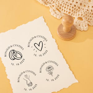 May include: A wooden stamp with a round handle is next to a sheet of paper with four different stamp designs. The stamps include the names "Ronald & Cynthia" and the date "10.16.2025" with images of wedding rings, a heart, holding hands, and a flower.