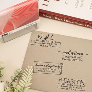 Puede incluir: Un sello de tinta rojo y blanco etiquetado "Printer 40" junto a un sobre beige con varios sellos de dirección negros. Los sellos incluyen nombres, direcciones y detalles florales. Un libro está en el fondo.