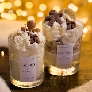 May include: Two glass candles with whipped cream topping, chocolate accents, and a gingerbread man. The candles have labels that read "cookies & cream." The background features blurred golden lights, creating a warm, festive ambiance.