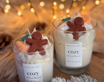 Puede incluir: Dos velas en vaso de cristal transparente con cera blanca, decoradas con cera batida, una figura de hombre de jengibre marrón y pequeños detalles naranjas y verdes. Cada vela lleva una etiqueta blanca con el texto "cozy christmas".