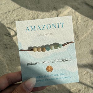 Könnte beinhalten: Ein Armband mit sieben kleinen, runden, mehrfarbigen Perlen an einem dünnen braunen Band, befestigt auf einer weißen Präsentationskarte. Die Karte zeigt den Text AMAZONIT, NATURSTEIN, Balance, Mut, Leichtigkeit sowie weiteren deutschen Text.