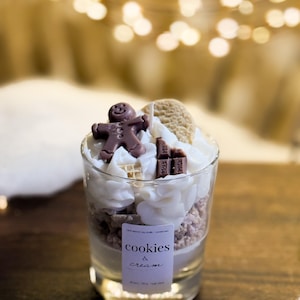 May include: A glass candle with a layered design of cookie crumbles, whipped cream, and chocolate decorations, including a gingerbread man. The candle is labelled "cookies & cream".