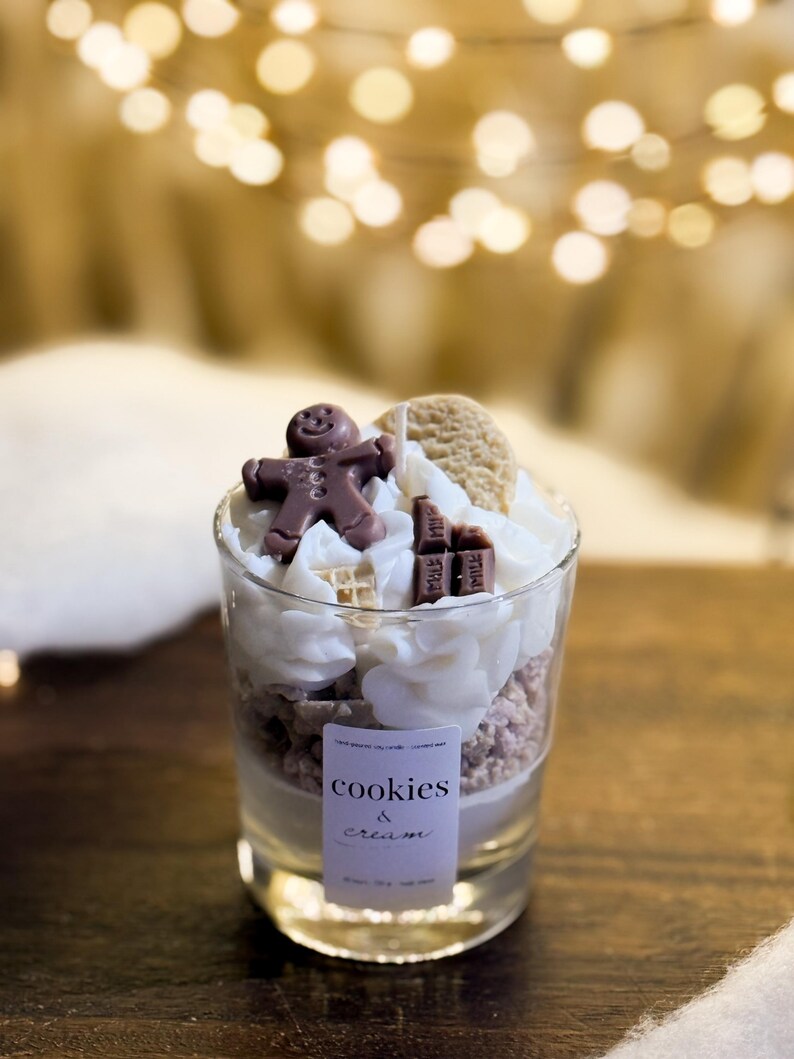 May include: A glass candle with a layered design of cookie crumbles, whipped cream, and chocolate decorations, including a gingerbread man. The candle is labelled "cookies & cream".