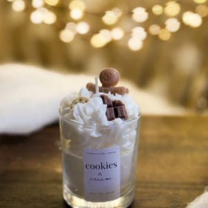 May include: A Cookies & Cream scented candle in a clear glass. The candle is decorated with white wax swirls, a gingerbread man, and chocolate pieces. The label reads "cookies & cream".