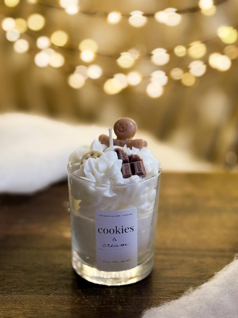 May include: A Cookies & Cream scented candle in a clear glass. The candle is decorated with white wax swirls, a gingerbread man, and chocolate pieces. The label reads "cookies & cream".