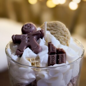 May include: A glass candle with a gingerbread man, a biscuit, and chocolate bars on top of white wax. The candle has a wick and is surrounded by fluffy white wax. The base of the candle is a light purple colour.