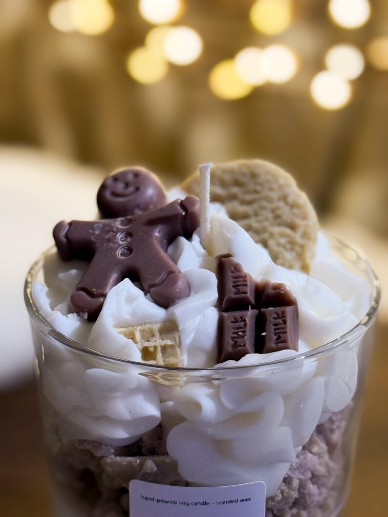 May include: A glass candle with a gingerbread man, a biscuit, and chocolate bars on top of white wax. The candle has a wick and is surrounded by fluffy white wax. The base of the candle is a light purple colour.