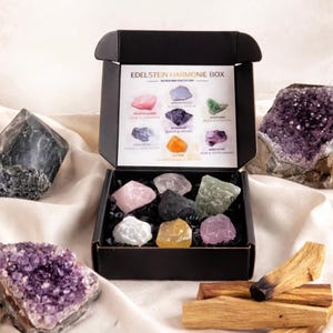 May include: A black box labelled "Edelstein Harmonie Box" containing various crystals, including rose quartz, amethyst, and citrine. The box is open, revealing the crystals inside. Additional crystals and several sticks of palo santo surround the box.