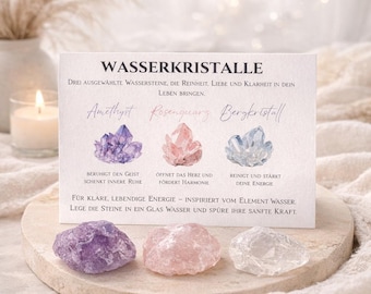 Könnte beinhalten: Eine rechteckige weiße Karte mit der Überschrift WASSERKRISTALLE, die Illustrationen von Amethyst, Rosenquarz und Bergkristall sowie deutschen Text zeigt. Drei rohe Steine in Violett, Rosa und Transparent liegen auf einem runden Steinsockel.