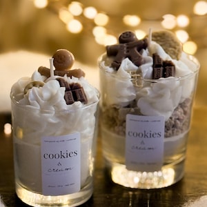 May include: Two glass candles with cookie-like layers, topped with whipped cream, chocolate pieces, and gingerbread men. Each candle has a label that reads "cookies & cream". The background features soft, blurred lights.