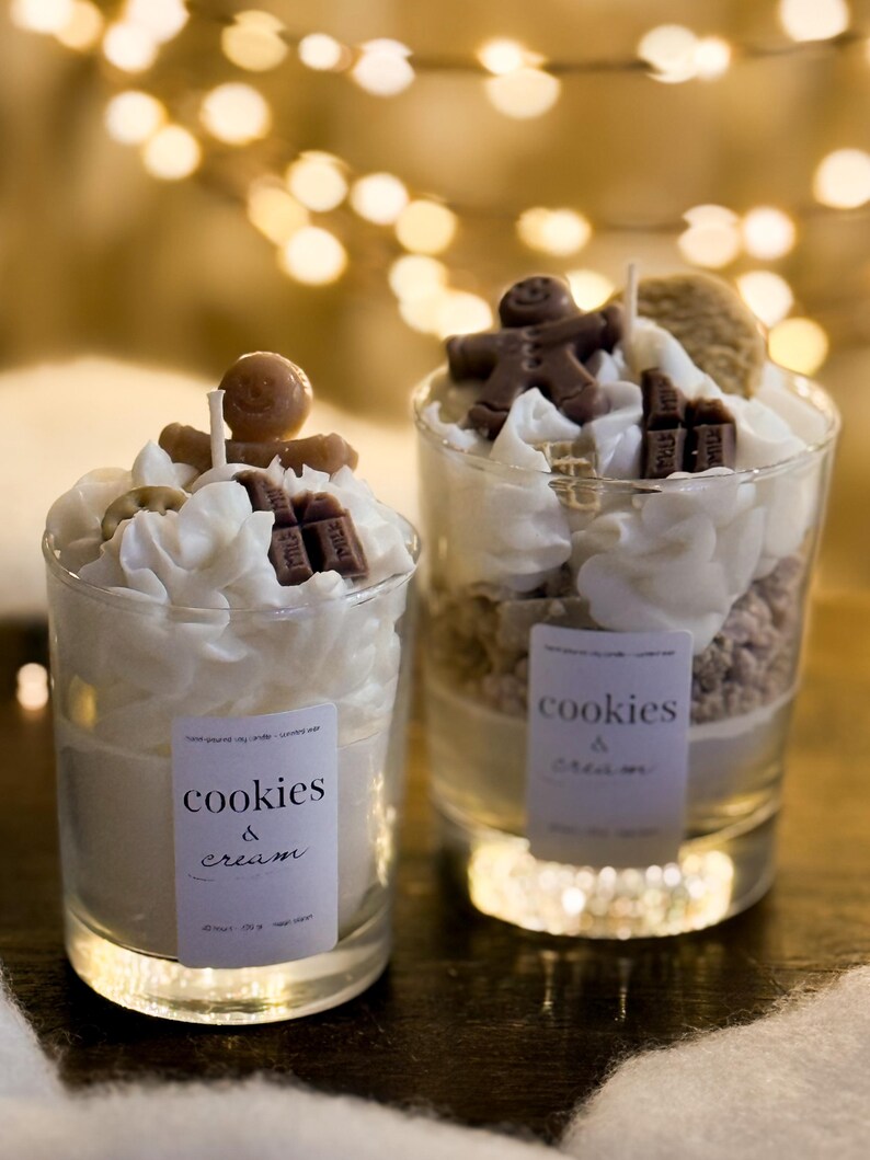 May include: Two glass candles with cookie-like layers, topped with whipped cream, chocolate pieces, and gingerbread men. Each candle has a label that reads "cookies & cream". The background features soft, blurred lights.