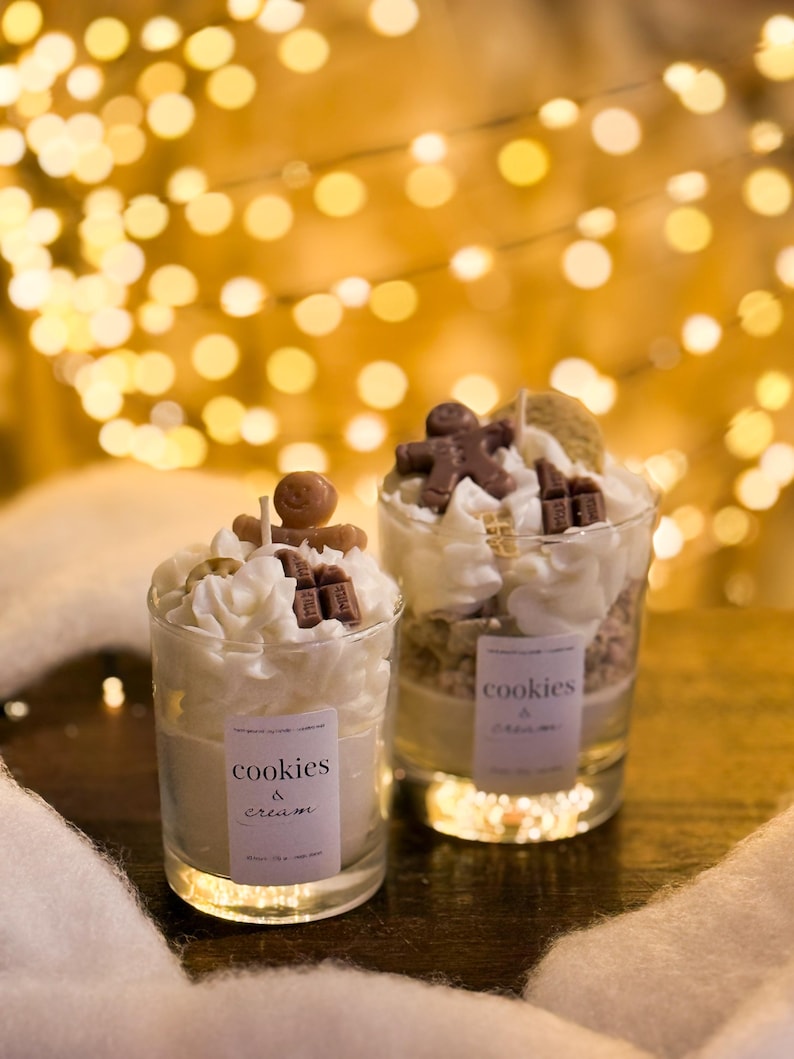 May include: Two glass candles with whipped cream topping, chocolate accents, and a gingerbread man. The candles have labels that read "cookies & cream." The background features blurred golden lights, creating a warm, festive ambiance.