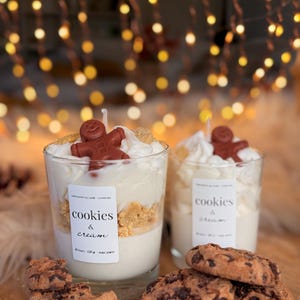 May include: Two glass candles topped with white wax swirls, brown gingerbread man shapes, and cookie crumbles. Each candle features a white label reading cookies & cream, 40 hours, 230 g, magic planet. Three chocolate chip cookies sit in the foreground.