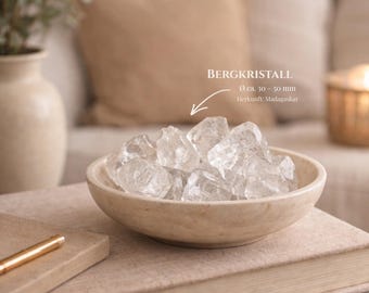 Könnte beinhalten: Eine beige Schale mit klaren Bergkristallen, ca. 3-5 cm groß, steht auf einem Stapel Bücher. Die Kristalle sind mit "Bergkristall" und dem Herkunftsort Madagaskar beschriftet. Ein goldener Stift liegt auf den Büchern.