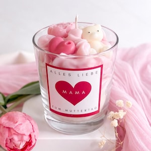 Op de afbeelding: Een glazen kandelaar gevuld met roze en witte wasvormen, waaronder harten en een teddybeer. De kaars heeft een wit etiket met een rood hart en het woord "MAMA". De tekst "Alles Liebe Zum Muttertag" is ook zichtbaar.