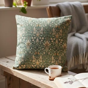 May include: Square decorative pillow with a green and gold floral pattern. The pillow is resting on a wooden bench next to a cup of tea and an open book. The pillow has a soft, inviting appearance.