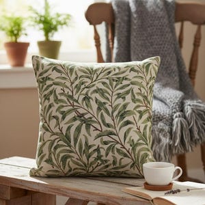 May include: Square decorative pillow with a green and brown leaf pattern. The pillow is on a wooden bench with a cup of tea and an open book. A gray blanket is draped over a wooden chair in the background.