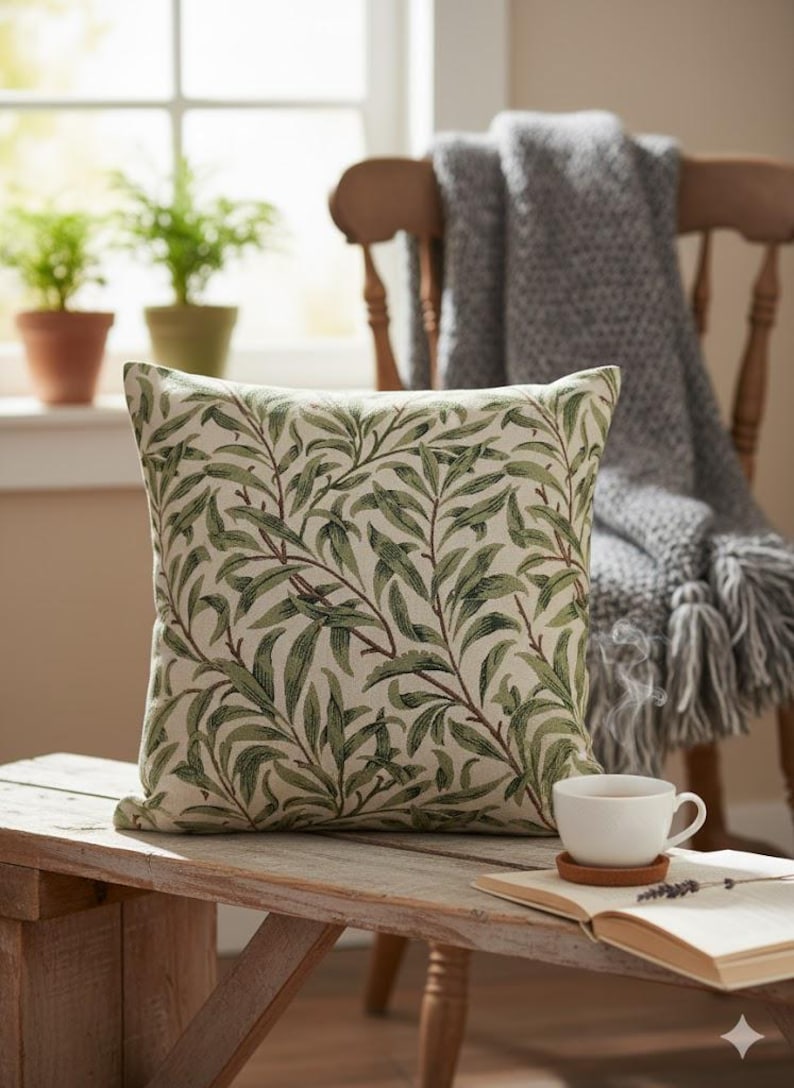 May include: Square decorative pillow with a green and brown leaf pattern. The pillow is on a wooden bench with a cup of tea and an open book. A gray blanket is draped over a wooden chair in the background.