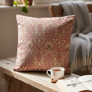 May include: A decorative square pillow with a red and gold floral pattern. The pillow is placed on a wooden bench next to a cup of tea and an open book. The pillow's design features intricate botanical details.