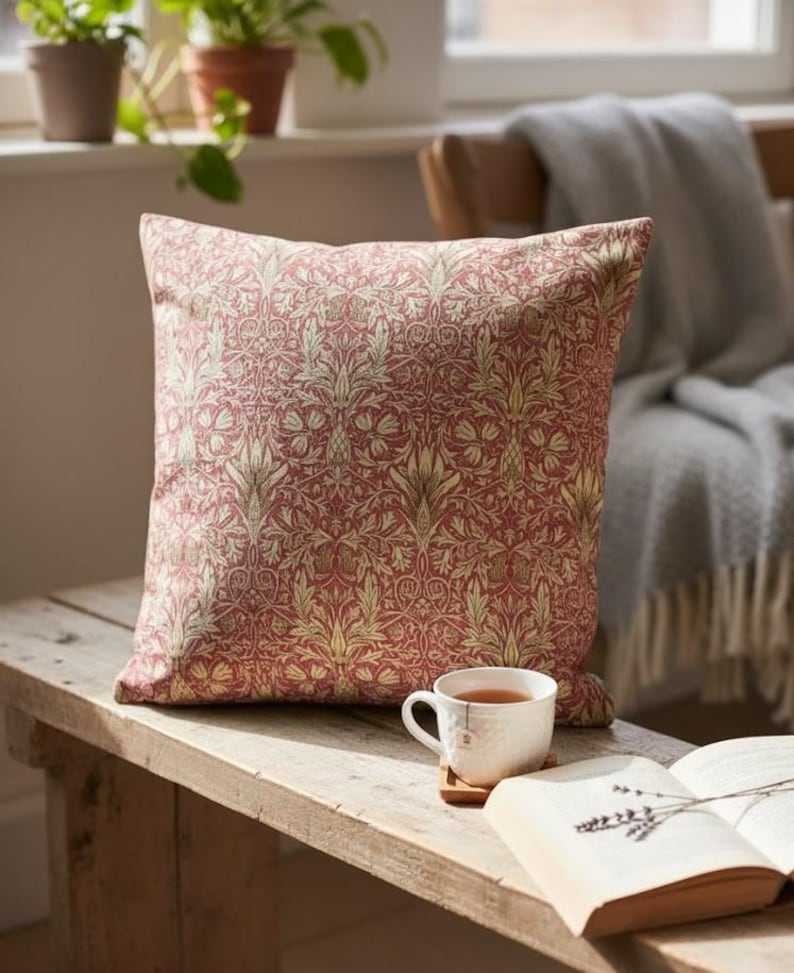 May include: A decorative square pillow with a red and gold floral pattern. The pillow is placed on a wooden bench next to a cup of tea and an open book. The pillow's design features intricate botanical details.