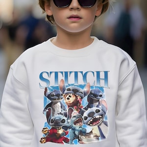 Op de afbeelding: Witte sweatshirt met een afbeelding van Stitch en andere personages uit de animatiefilm. Het woord "STITCH" is in blauw boven de personages gedrukt. De sweatshirt wordt gedragen door een persoon met een zonnebril.
