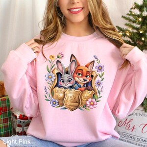 Puede incluir: Sudadera rosa claro con un dise&ntilde;o colorido de Judy Hopps y Nick Wilde de Zootopia, rodeados de flores y escudos de polic&iacute;a. Las palabras "ZOOTO" y "POLICE" son visibles en los escudos. La sudadera es de cuello redondo.