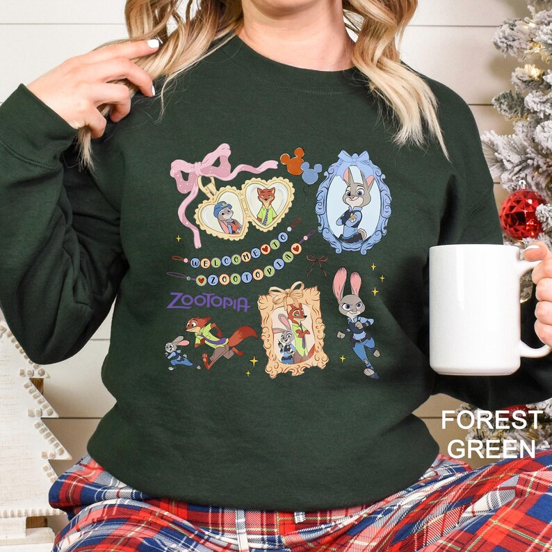 May include: Forest green sweatshirt featuring Zootopia characters in framed illustrations. The design includes Judy Hopps and Nick Wilde, with the word "Zootopia" in a stylized font. A white mug is held in the hand.