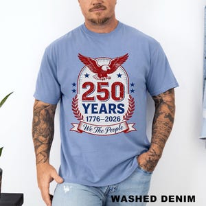 May include: Light blue t-shirt featuring a patriotic design. The graphic includes a red eagle, the number "250" in a bold font, and the text "YEARS 1776-2026 We The People". The shirt is made of a soft material and has short sleeves.