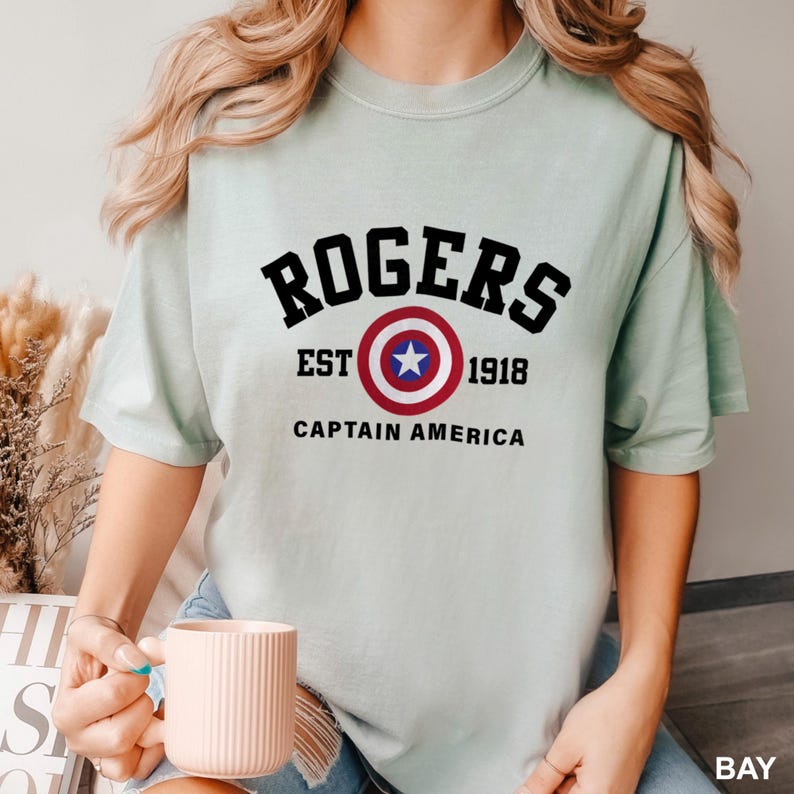 May include: A light blue t-shirt with a black graphic of a Captain America shield and the text "ROGERS EST 1918 CAPTAIN AMERICA".