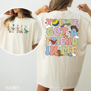 May include: Ivory t-shirt with a colourful Toy Story character graphic on the front and the phrase "You've Got a Friend in Me" on the back. The design includes a rocket, cowboy hat, and other playful elements.