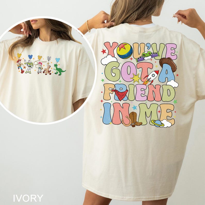 May include: Ivory t-shirt with a colourful Toy Story character graphic on the front and the phrase "You've Got a Friend in Me" on the back. The design includes a rocket, cowboy hat, and other playful elements.