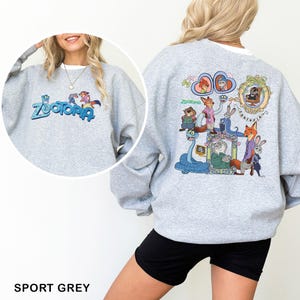 K&ouml;nnte beinhalten: Sportgraues Sweatshirt mit dem Wort "Zootopia" in Blau und T&uuml;rkis auf der Vorderseite. Auf der R&uuml;ckseite des Sweatshirts befindet sich eine farbenfrohe Grafik von Charakteren aus dem Animationsfilm "Zootopia".