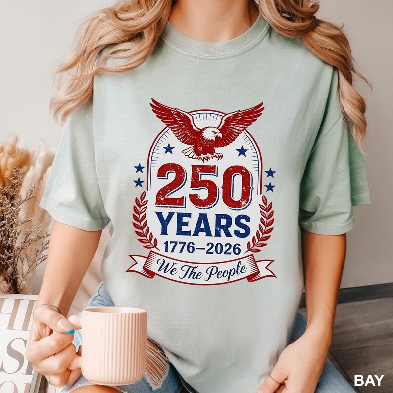 May include: A light green t-shirt featuring a patriotic design. The design includes a red eagle, the text "250 YEARS 1776-2026", and the phrase "We The People". The shirt is a soft, comfortable material.