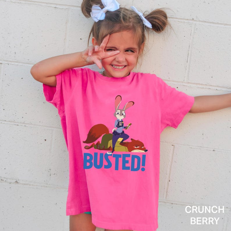 May include: A bright pink t-shirt featuring a cartoon image of a rabbit and a fox with the word "BUSTED!" in blue. The shirt is a solid color and has short sleeves.