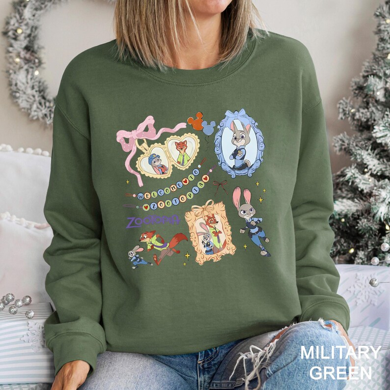 May include: Military green sweatshirt featuring framed cartoon characters from Zootopia. The design includes Judy Hopps and Nick Wilde, with the word "Zootopia" in purple. The sweatshirt has a crew neck and long sleeves.