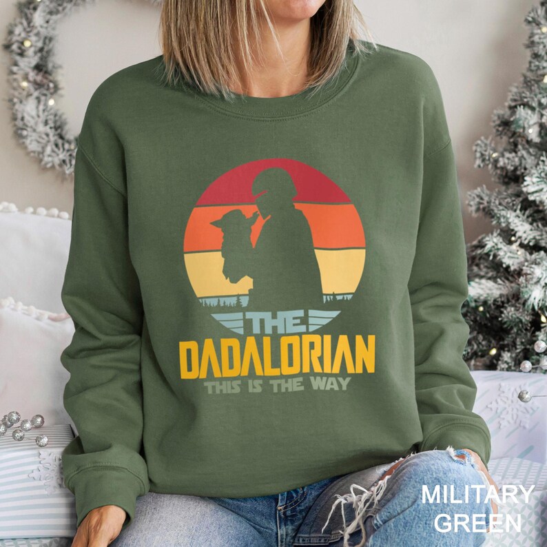 May include: A military green crewneck sweatshirt featuring a graphic of a silhouette of a helmeted figure holding a small creature against a sunset background. The text reads THE DADALORIAN THIS IS THE WAY in yellow and white lettering.