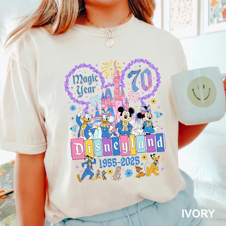 May include: Ivory-colored t-shirt featuring a colorful Disneyland graphic. The design includes the text "Magic Year" and "70", along with Disney characters and the Disneyland castle. The text "Disneyland 1955-2025" is also present.