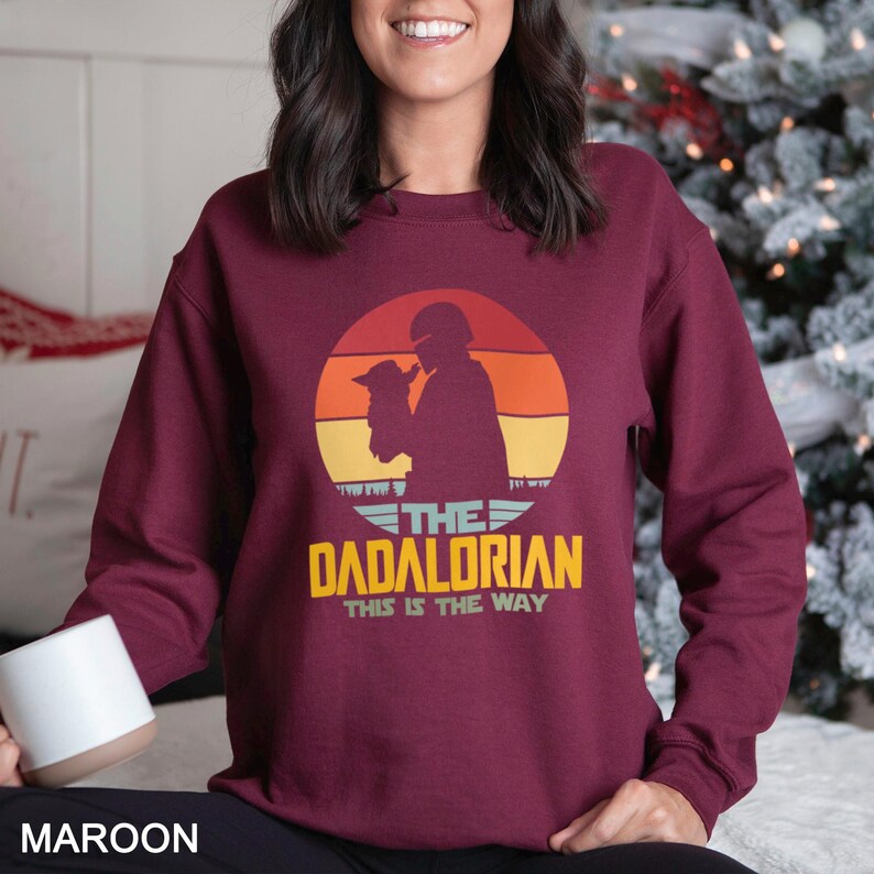 May include: A maroon long-sleeve sweatshirt featuring a graphic of a silhouette of a helmeted figure holding a small creature against a sunset background. The text reads THE DADALORIAN THIS IS THE WAY in yellow and white lettering on the front.
