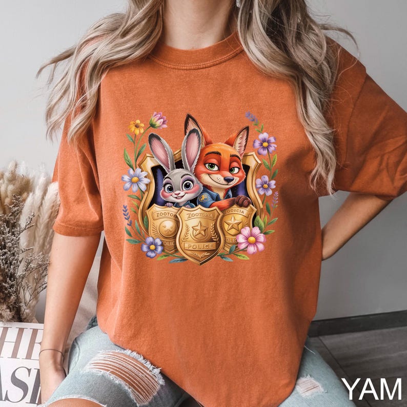 May include: Burnt orange t-shirt with a colourful graphic of Judy Hopps and Nick Wilde from Zootopia, framed by flowers and police badges. The shirt has a relaxed fit and short sleeves, suitable for casual wear.