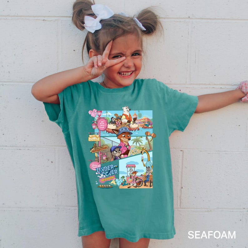 May include: A teal t-shirt with a vibrant cartoon graphic showing animal characters in a theme park, with the words "UNDER SCUER MISSION". The t-shirt is worn by a young girl, who is smiling and giving a peace sign.