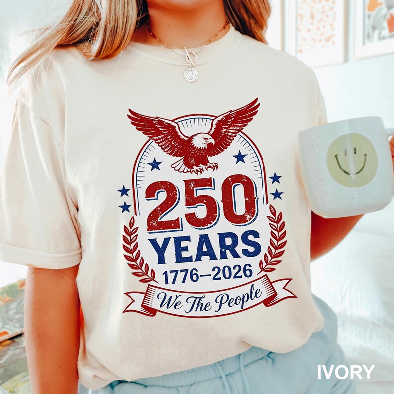 May include: Ivory-colored t-shirt featuring a patriotic design. The graphic includes a red eagle with outstretched wings, the text "250 YEARS 1776-2026 We The People", and stars. A white mug with a smiley face is held in the image.