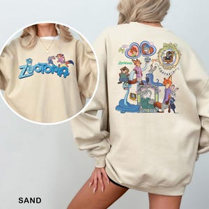 K&ouml;nnte beinhalten: Sandfarbener Pullover mit dem Wort "Zootopia" in Blau und einer farbenfrohen Grafik von Charakteren aus dem Animationsfilm. Auf der R&uuml;ckseite des Sweatshirts befindet sich eine gr&ouml;&szlig;ere, detaillierte Illustration der Zootopia-Charaktere. Das Wort "SAND" ist unten aufgedruckt.