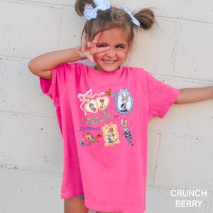 May include: A bright pink t-shirt featuring multiple framed images of characters from the animated movie "Zootopia". The shirt has the word "Zootopia" printed in a stylized font. The text "CRUNCH BERRY" is printed in the bottom right corner.